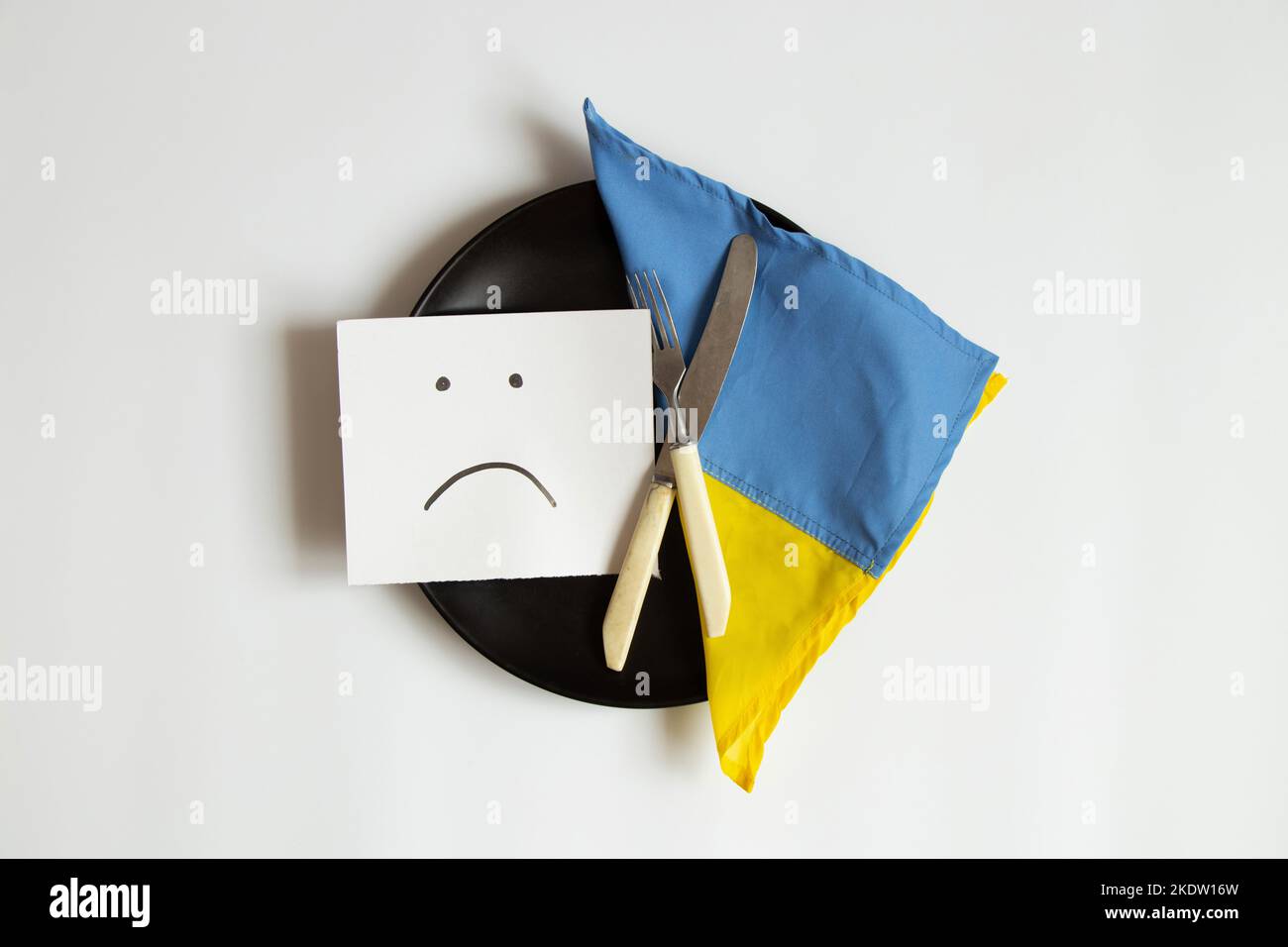 Un triste emoticon è disegnato sulla carta, si trova su un piatto accanto alla bandiera dell'Ucraina su uno sfondo bianco, tristezza e mancanza di cibo, Ucraina senza cibo e. Foto Stock