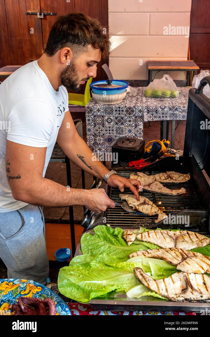 Un giovane uomo cucinare il pesce su Una griglia al Ballaro Street Market, Palermo, Sicilia, Italia. Foto Stock