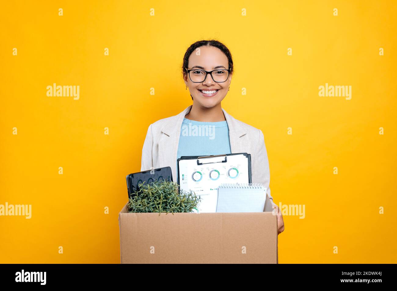 Nuovo lavoro. Eccitato positivo bella gara mista femminile impiegato, nuovo arrivato, in abiti eleganti, tenendo scatola di cartone, stand su sfondo isolato arancione, guardando la fotocamera, sorridendo amichevole Foto Stock