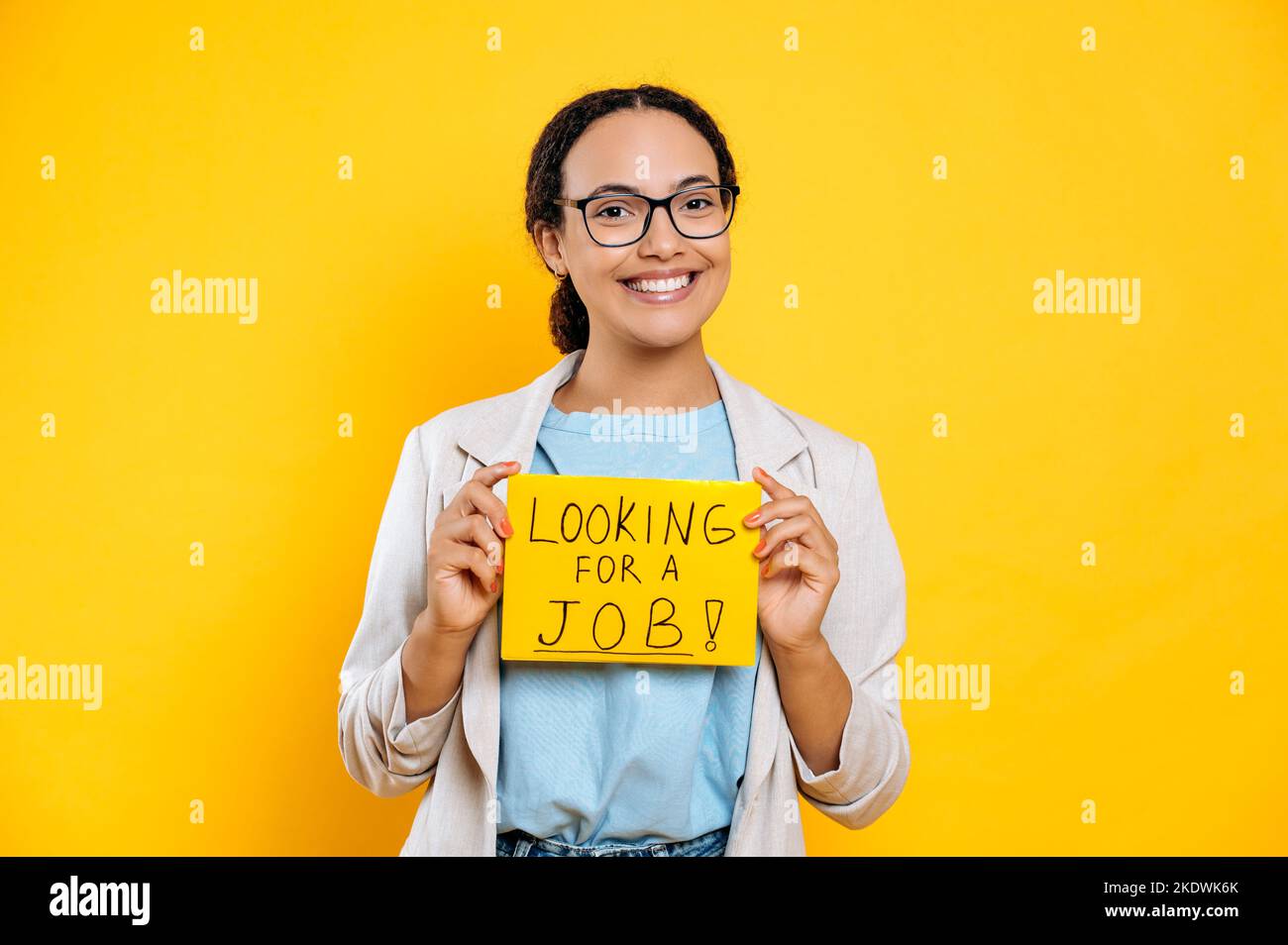 Eccitato positivo successo brasiliana o ispanica giovane donna, specialista creativo alla ricerca di un lavoro, si trova su sfondo isolato arancione, mostra un segno alla ricerca di un lavoro, guarda la fotocamera, sorridendo Foto Stock