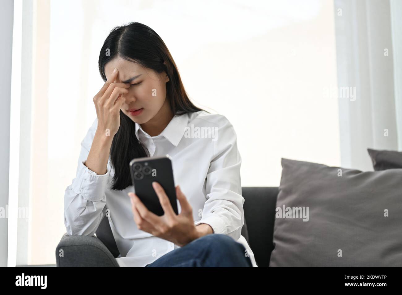 Stressato giovane asiatica utilizzando il suo smartphone mentre si siede sul divano, soffre di mal di testa o stress oculare, sensazione di turbamento o insoddisfazione con alcuni Foto Stock