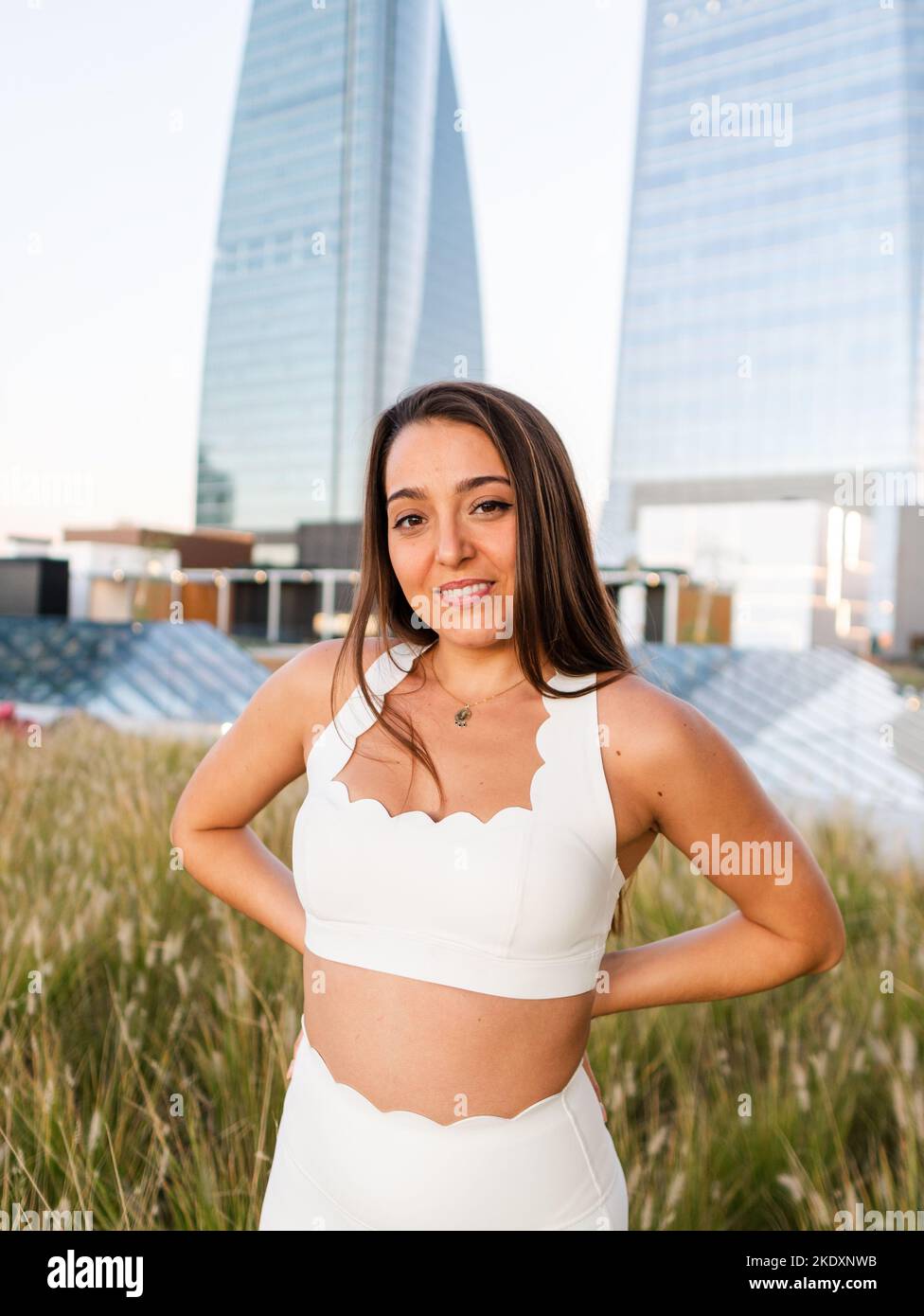 Giovane donna positiva con lunghi capelli castani in cima bianca e pantaloni in piedi sul prato con le mani dietro gli edifici moderni con pareti di vetro w Foto Stock
