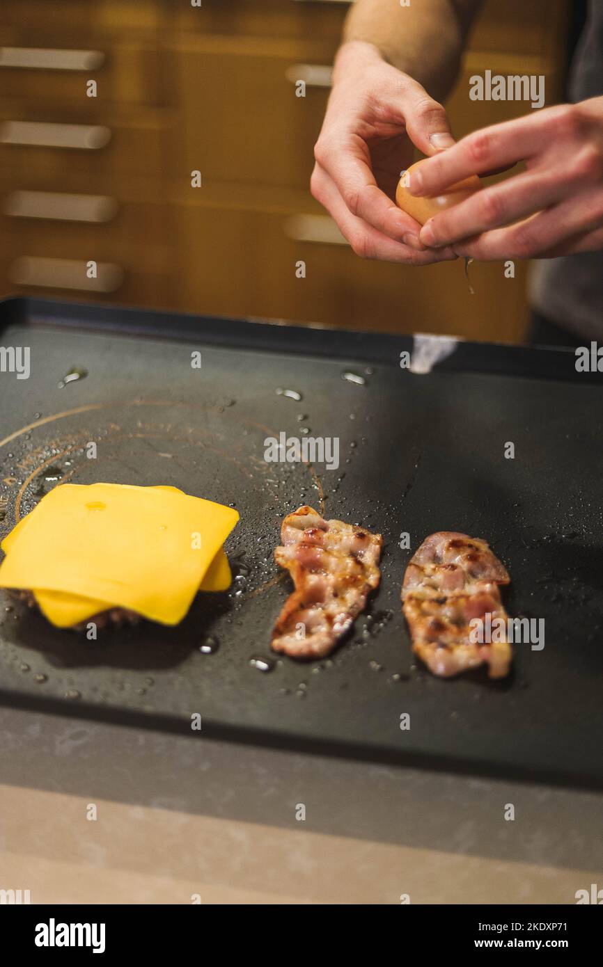 Angolo alto di raccolto anonimo cuoco rompere uova crude sulla griglia vicino al formaggio e fette di pancetta, mentre preparare delizioso hamburger in cucina Foto Stock
