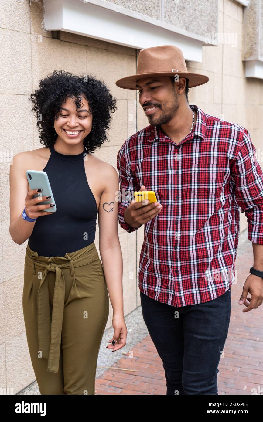 Allegra donna latino-americana in abiti casual che mostra video sul cellulare al ragazzo in camicia a scacchi e cappello, mentre camminando sulla strada della città in Wee Foto Stock