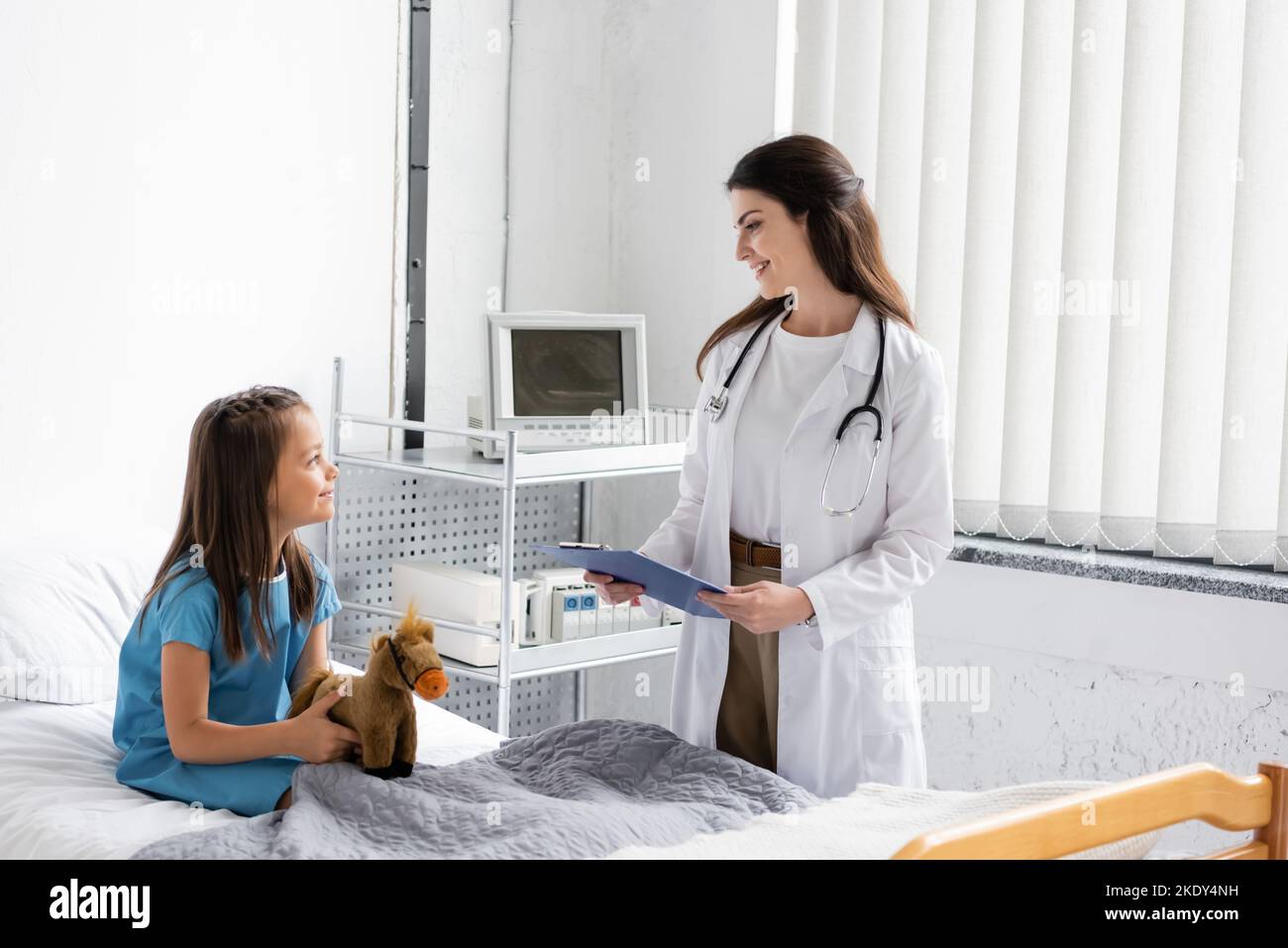Vista laterale di un medico sorridente con appunti che guarda il paziente con un giocattolo morbido in clinica, immagine stock Foto Stock
