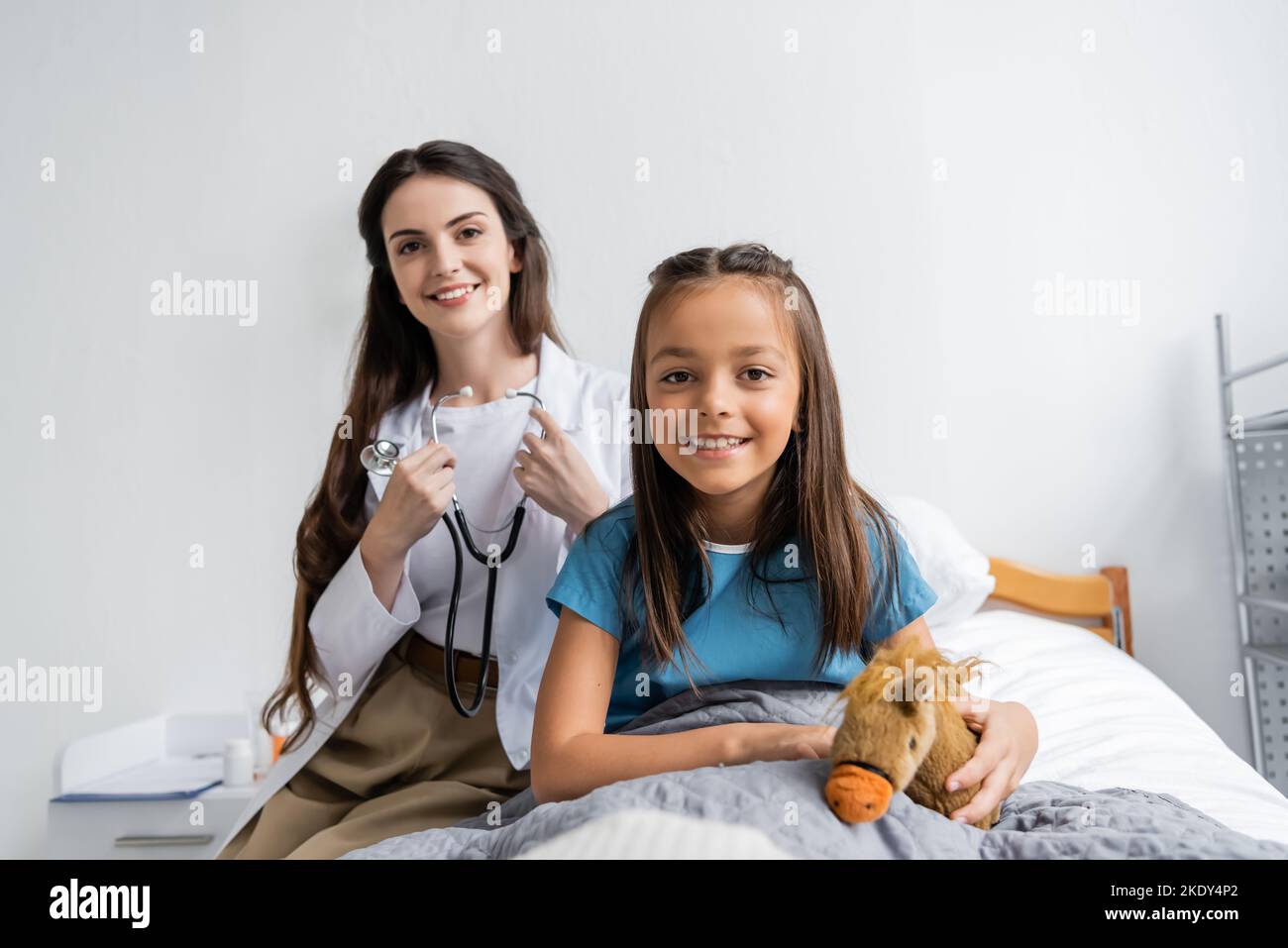Bambino sorridente che tiene il giocattolo molle vicino al medico con stetoscopio che guarda la macchina fotografica nel reparto dell'ospedale, immagine di scorta Foto Stock