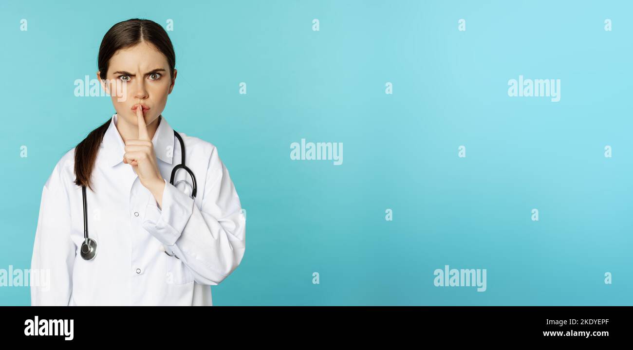 Medico donna arrabbiato, operatore sanitario che urla con disapprovazione, tabù silenzio gesto, silenzio qualcuno, in piedi su sfondo torchese Foto Stock