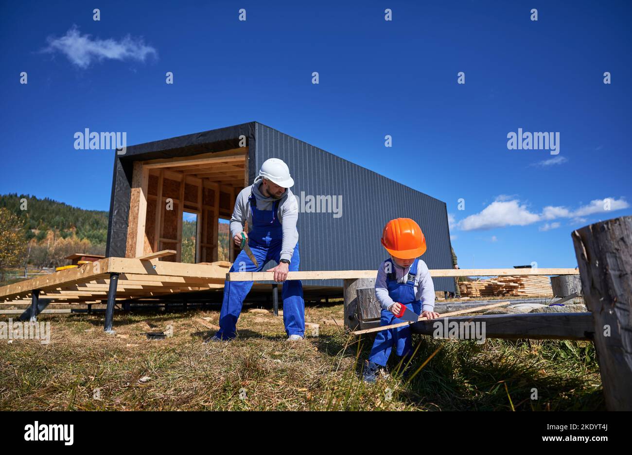 Padre con il figlio del bambino che costruisce la casa della struttura di legno sulla fondazione del palo. Costruttori maschi con sega a mano per tagliare tavole in cantiere nelle giornate di sole. Falegnameria e concetto di famiglia. Foto Stock