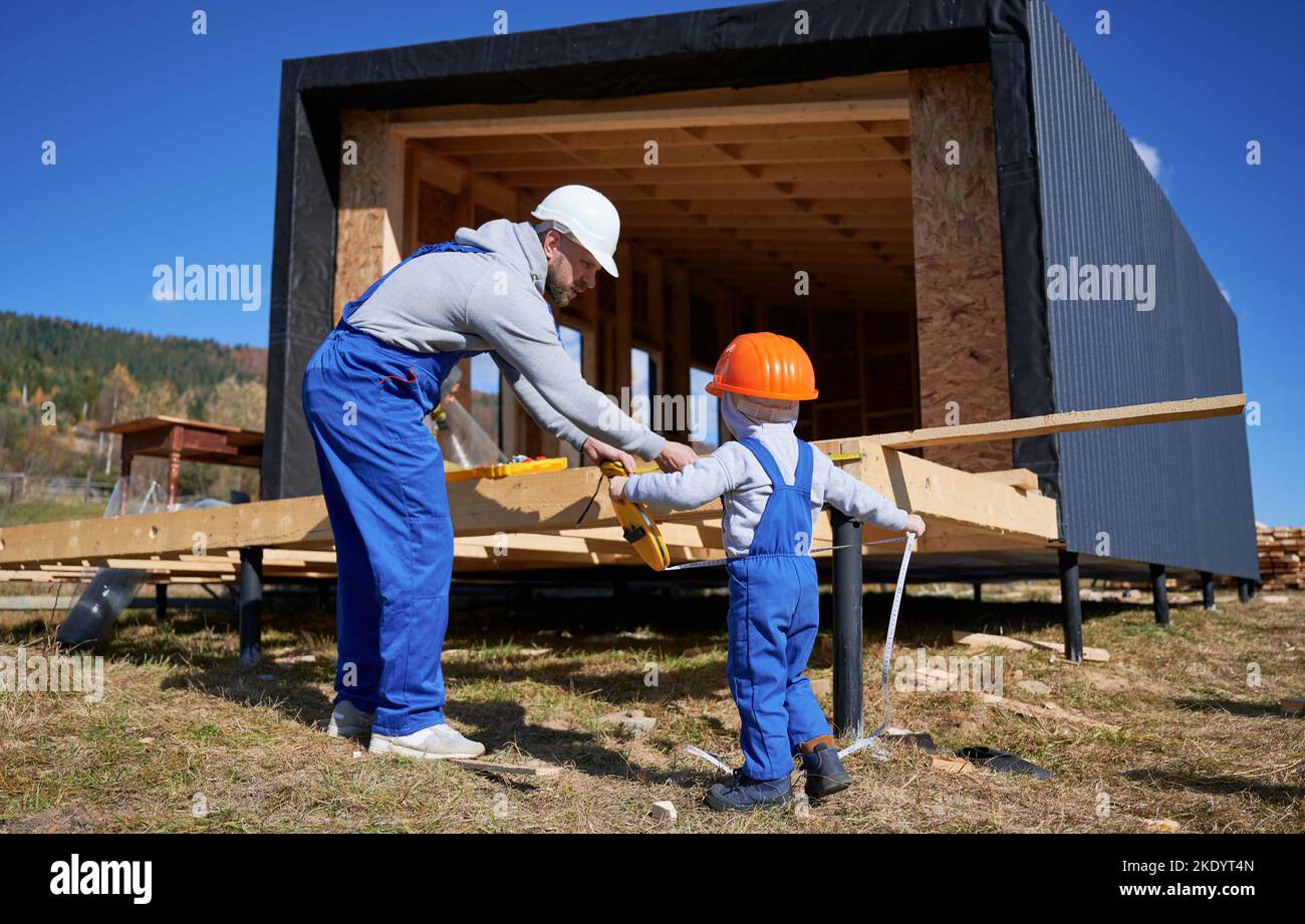 Padre con bambino figlio costruzione legno cornice casa. Costruttore maschile e bambino che gioca con metro a nastro sul cantiere, indossare casco e tute blu in giornata di sole. Carpenteria e concetto di famiglia. Foto Stock