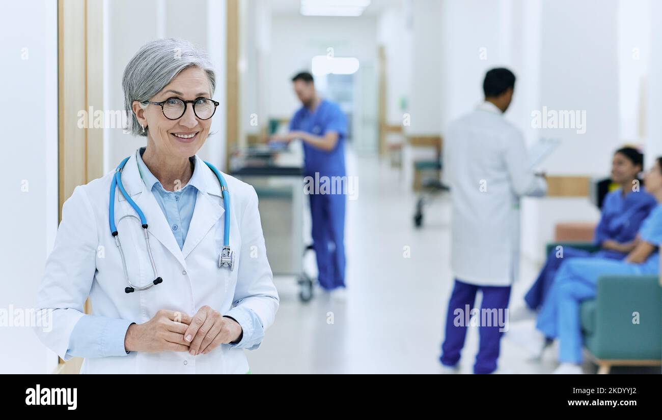 istituzione medica. Medico sorridente maturo in piedi nel corridoio di ospedale moderno sullo sfondo di assistenti medici e infermieri funzionanti Foto Stock