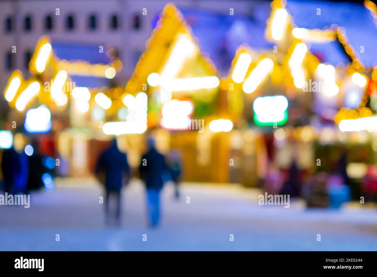 Sfondo sfocato. La gente cammina nella piazza della città la sera d'inverno. Silhouette nere di persone che camminano vicino a case decorate illuminazione luminosa. Luce bianca bokeh sfocare i punti dalle luci della casa incandescente Foto Stock