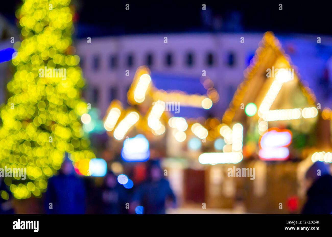 Sfondo sfocato. La gente cammina nella piazza della città la sera d'inverno. Silhouette nere di persone che camminano vicino a case decorate illuminazione luminosa. Luce bianca bokeh sfocare i punti dalle luci della casa incandescente Foto Stock