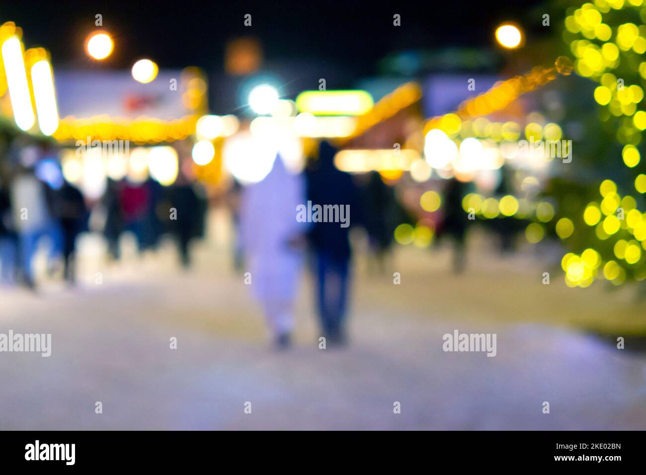 Sfondo sfocato. La gente cammina nella piazza della città la sera d'inverno. Silhouette nere di persone che camminano vicino a case decorate illuminazione luminosa. Luce bianca bokeh sfocare i punti dalle luci della casa incandescente Foto Stock