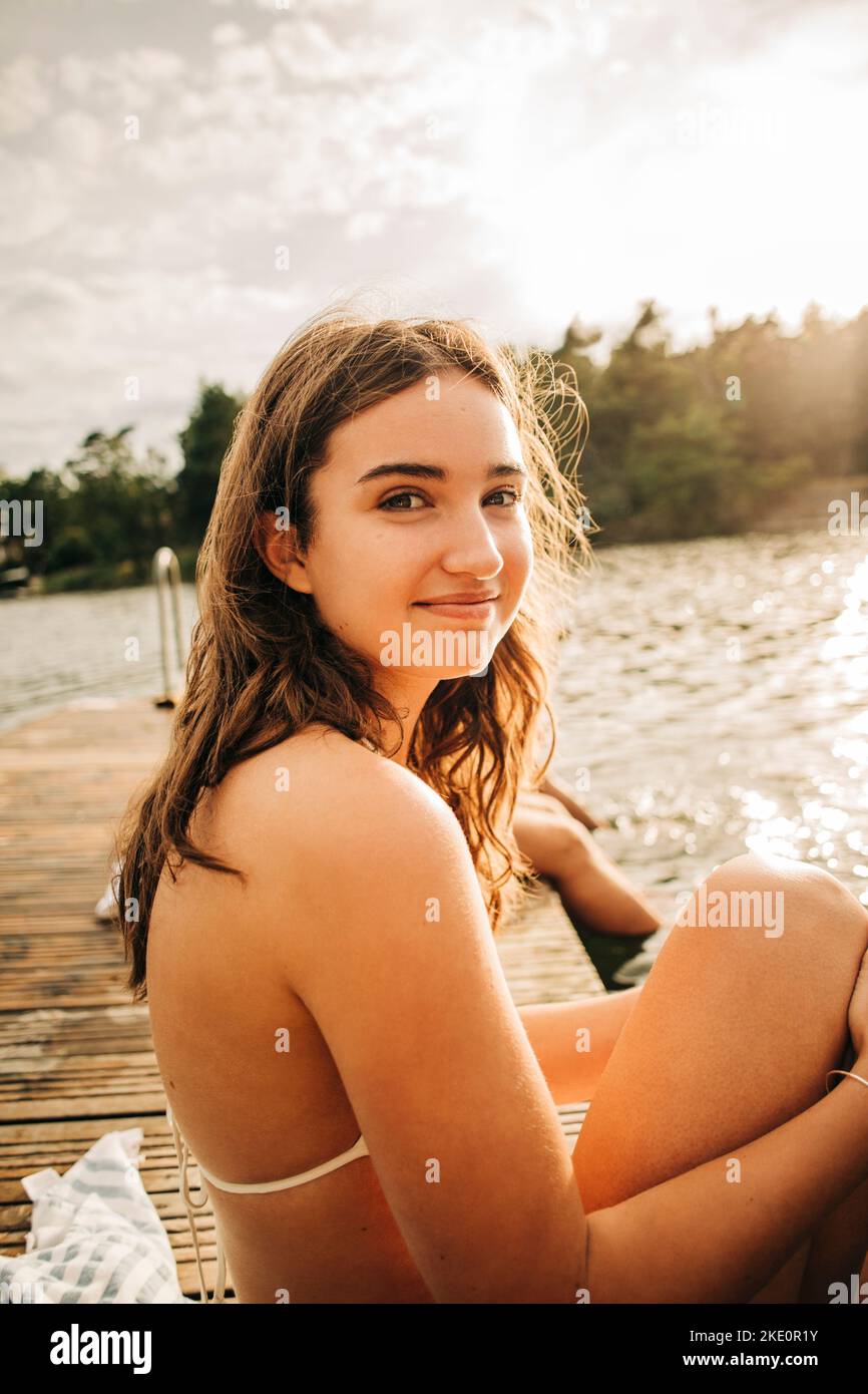 Vista laterale di una donna sorridente seduta sul molo sul lago durante le vacanze Foto Stock