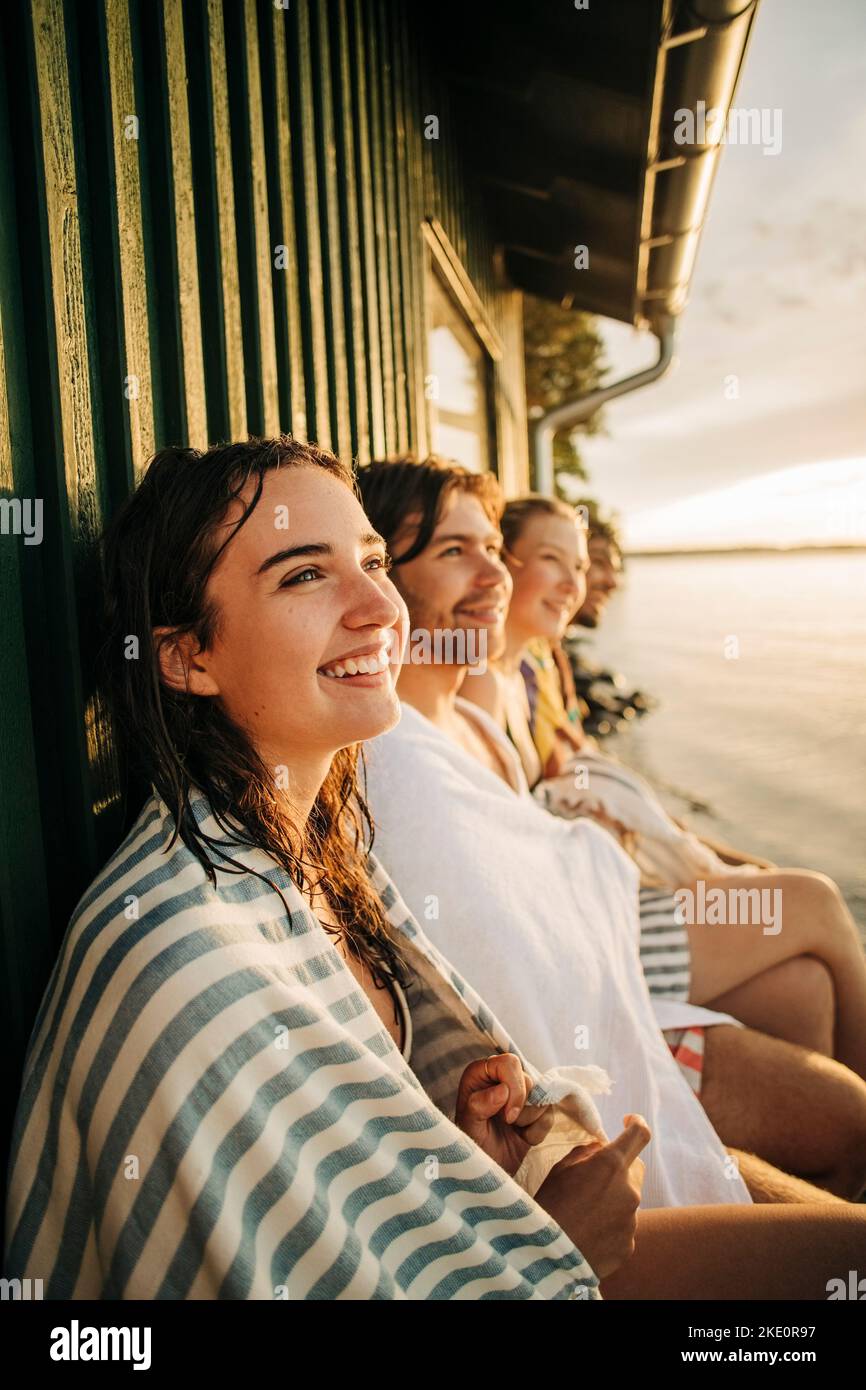 Felice donna contemplativa che indossa asciugamano seduto con gli amici vicino al cottage Foto Stock