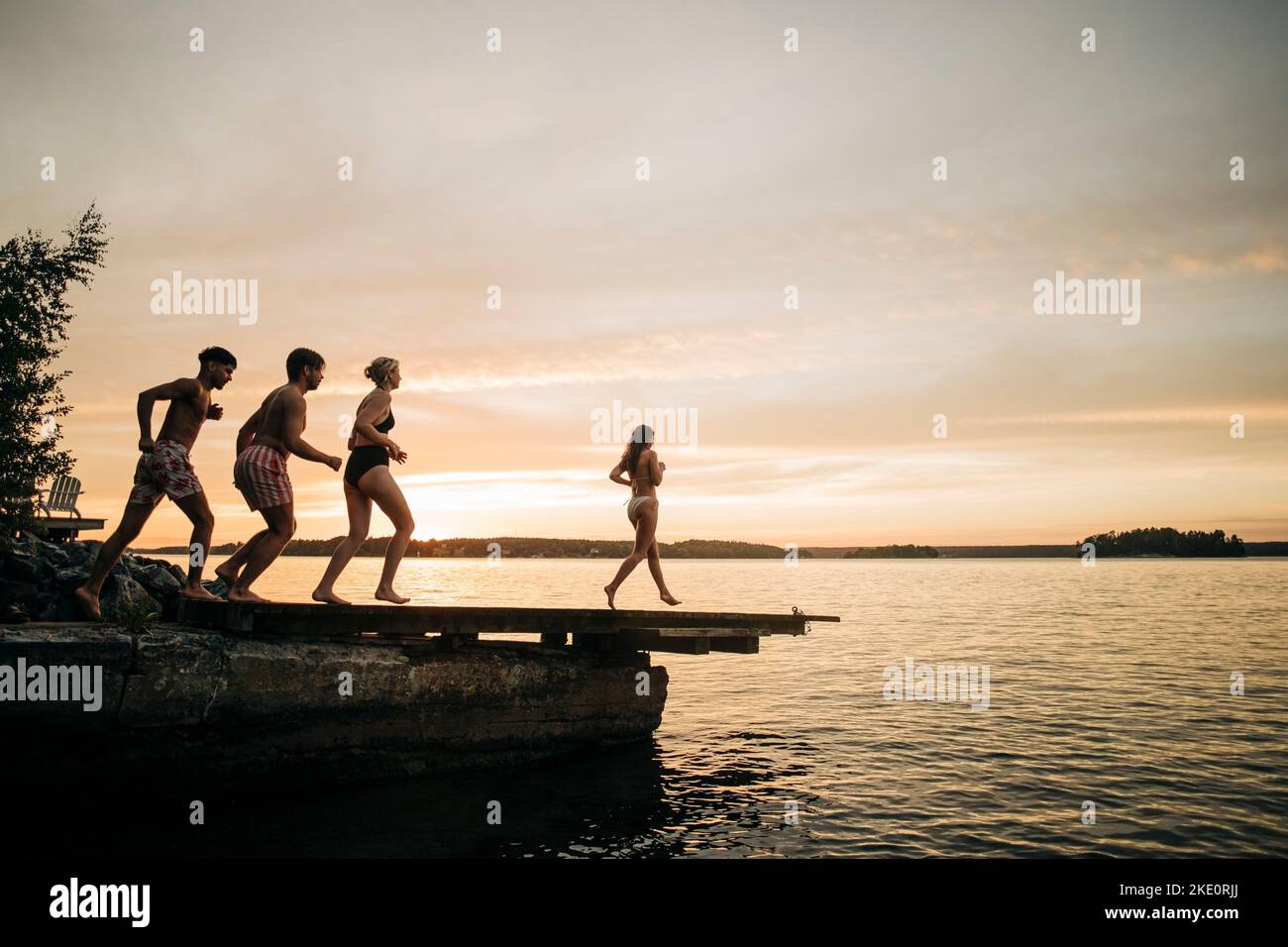 Amici maschili e femminili che corrono sul molo verso il lago durante le vacanze al tramonto Foto Stock