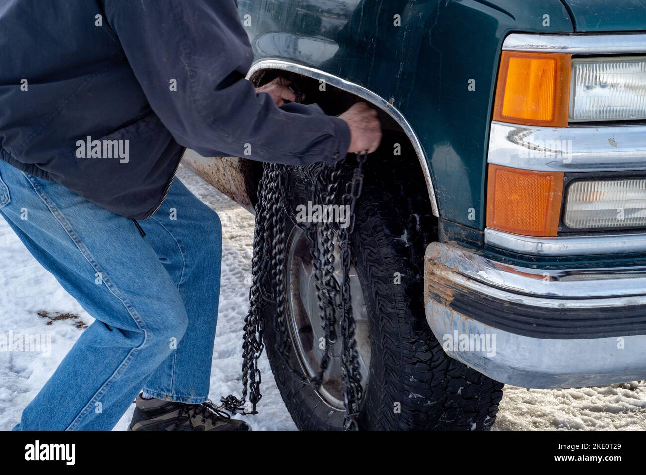 Un uomo anziano non identificabile, di 79 anni, solleva catene da neve sul pneumatico del suo pickup verde. Foto Stock