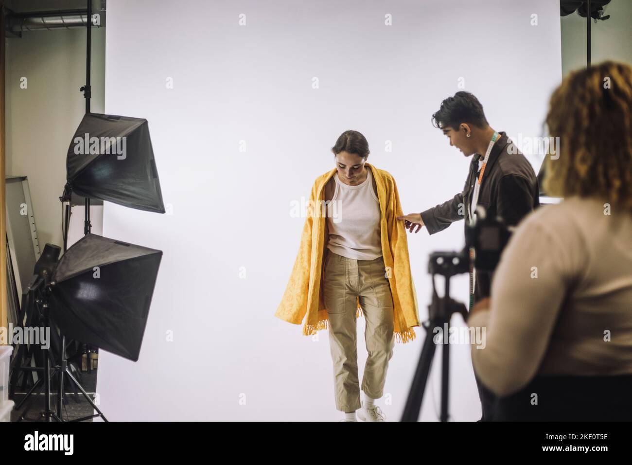 Designer maschile che assiste la modella femminile contro lo sfondo bianco durante le riprese fotografiche in studio Foto Stock
