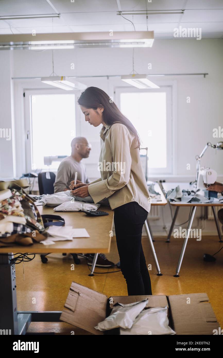 Vista laterale di una stilista di moda femminile che scrive sul pacchetto mentre lavora in officina Foto Stock