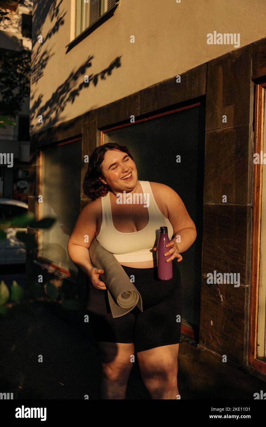 Giovane donna felice con il tappetino di esercitazione e la bottiglia dell'acqua in piedi nel cortile posteriore Foto Stock