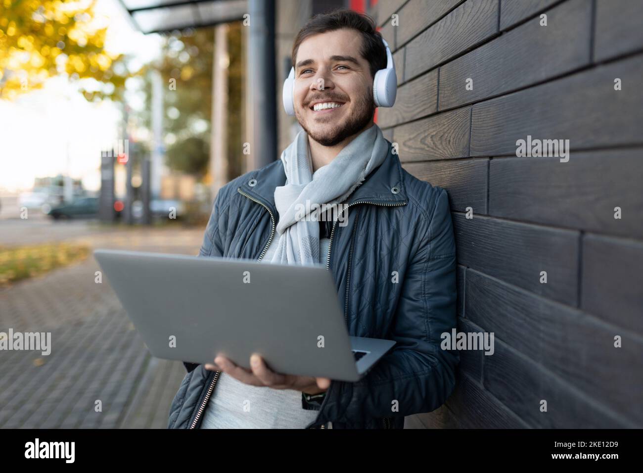 giovane uomo elegante che ascolta la musica con le cuffie e che gli copre gli occhi con un computer portatile nelle mani all'esterno Foto Stock
