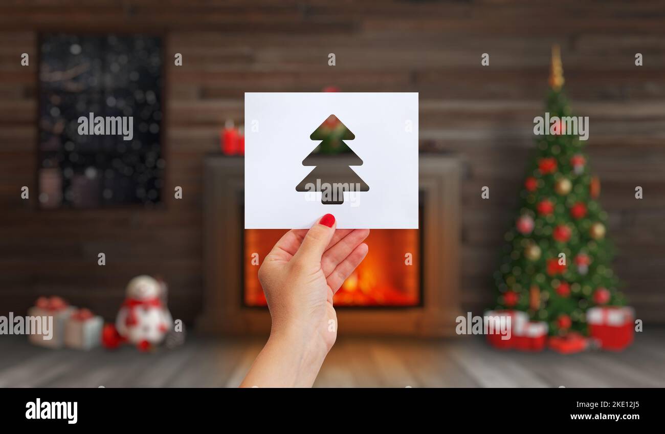La mano tiene una carta con un albero di Natale tagliato su di esso. Camino con un fuoco, un albero di Natale e decorazioni natalizie Foto Stock