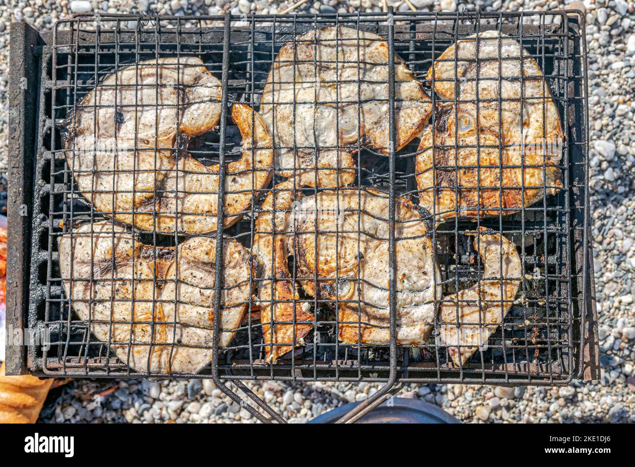 Vista dall'alto di fette di pesce spada grigliate in una griglia barbecue con un pane e ciottoli da spiaggia sfocati sullo sfondo. Foto Stock