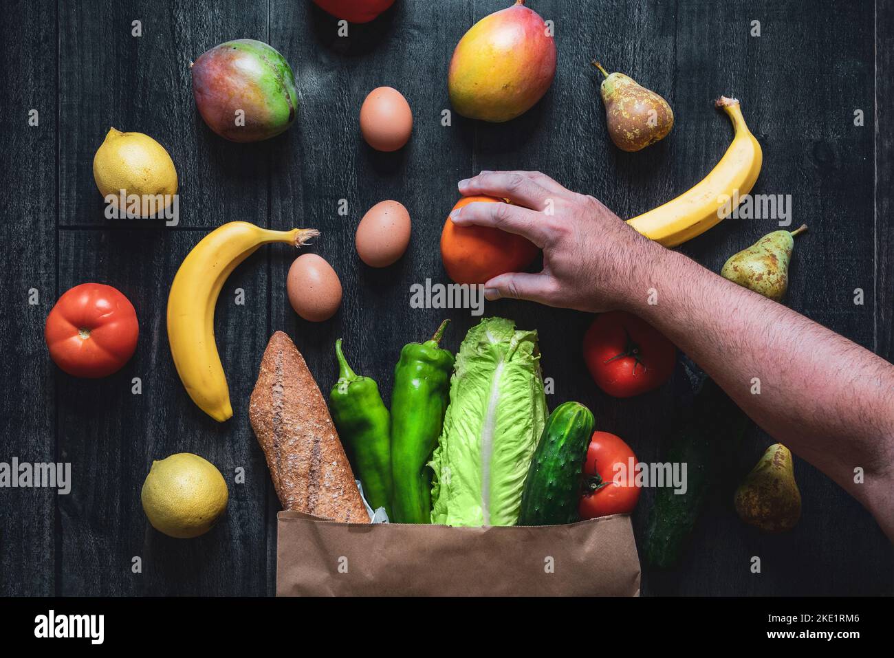 Una mano che raccoglie una frutta da un sacchetto di shopping riempito di verdure, frutta, uova e pane su un fondo di legno. Scelte alimentari sane. Superma online Foto Stock