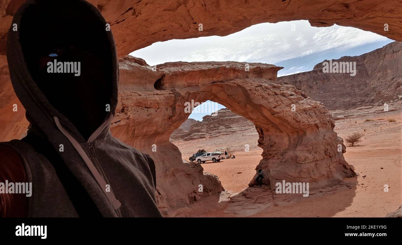 Selfie in una grotta del Sahara. Arco naturale, mesa, cielo nuvoloso, 4x4 automobili, uomini touareg in background. Un uomo irriconoscibile indossa un cappellino e il suo volto Foto Stock