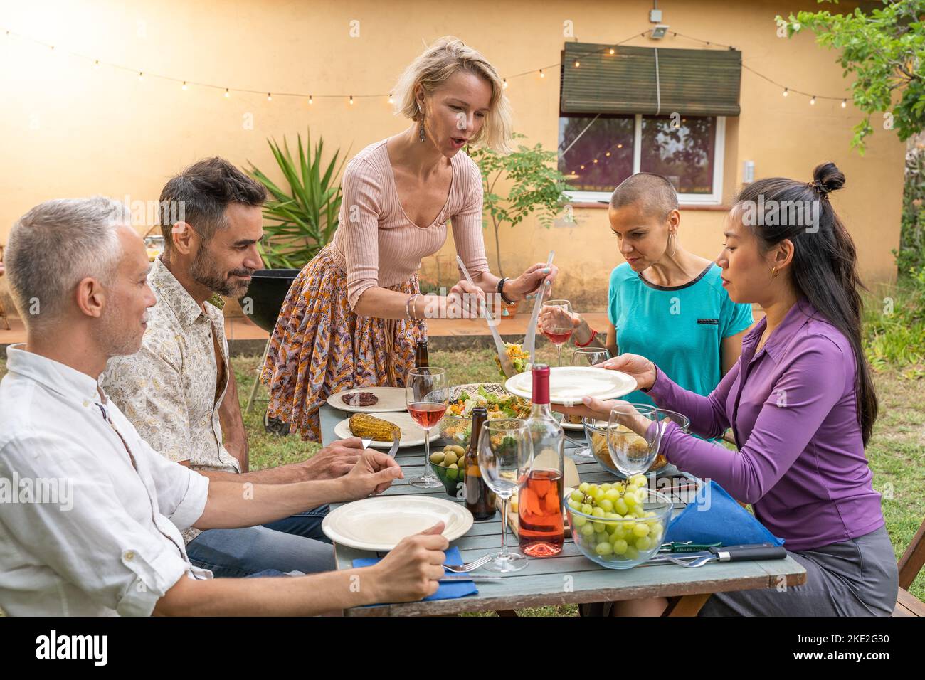 Amici multiculturali che ridono con un grande sorriso intorno al tavolo. Donna serve il cibo nel patio della casa. Sorrisi, concetto di allegria. Foto Stock