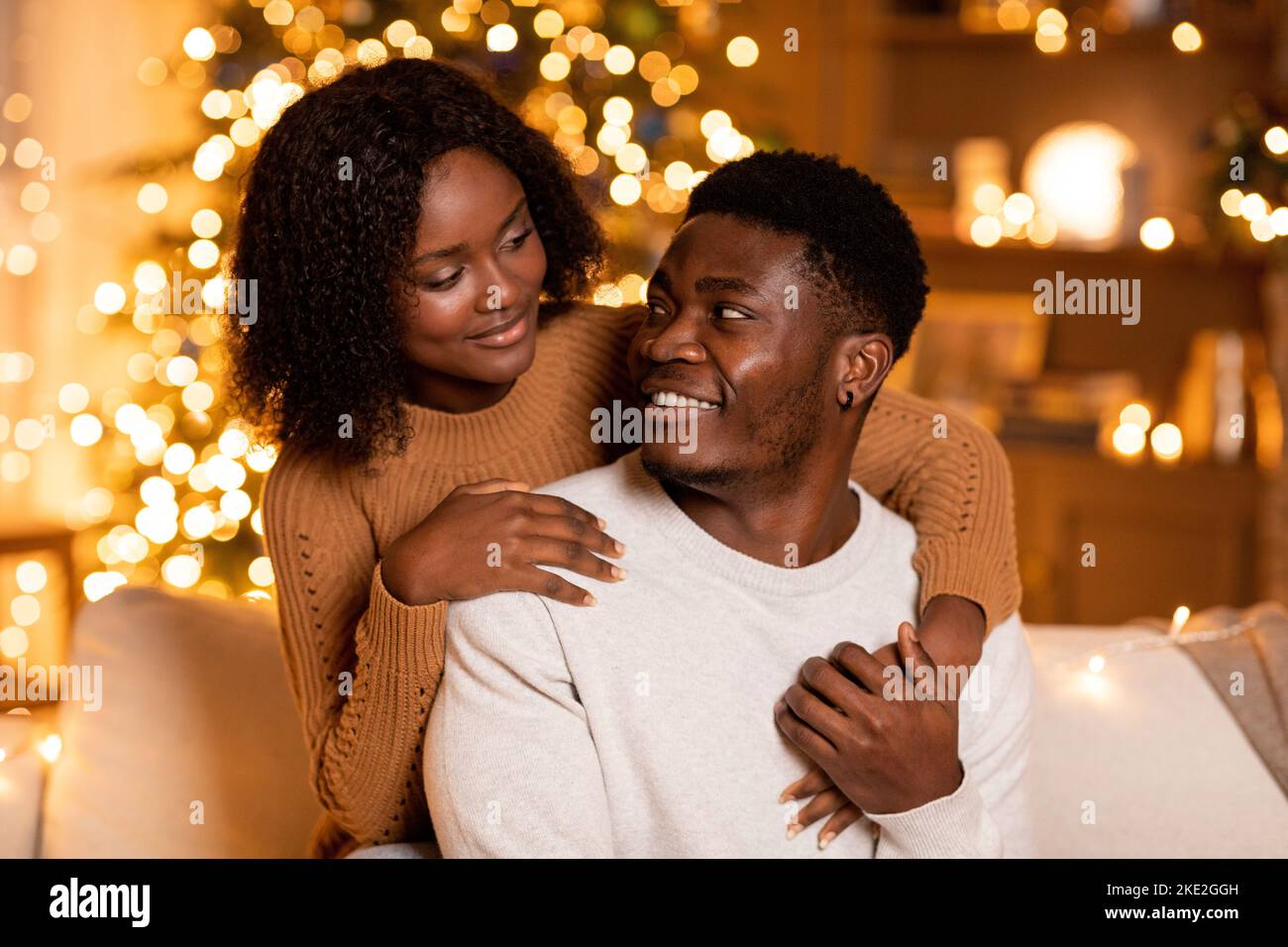Allegra donna afroamericana millenaria che abbraccia l'uomo, godersi le vacanze insieme nel soggiorno interno Foto Stock