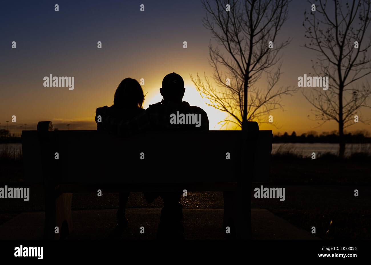 silhouette di una coppia eterosessuale che guarda il tramonto, seduto insieme su una panchina di fronte a un lago Foto Stock
