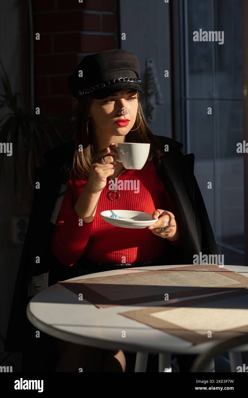 Bere caffè in una caffetteria. La ragazza di moda in un cappotto siede alla finestra di un caffè. Goditi il tempo libero Foto Stock