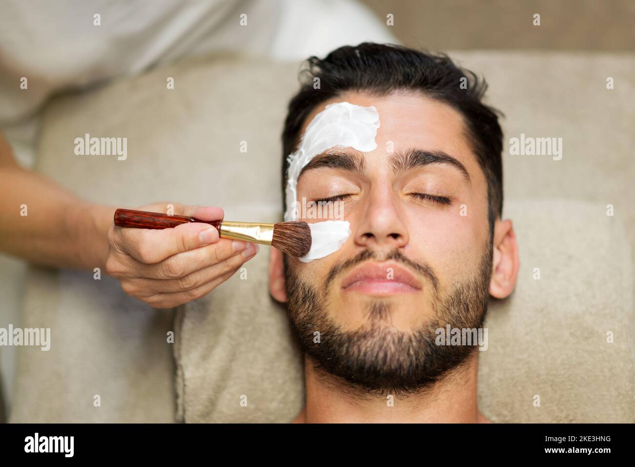 Dall'alto estetista raccolto applicando maschera di argilla sulla faccia di calmo bell'uomo bearded sdraiato sul tavolo in salone termale Foto Stock