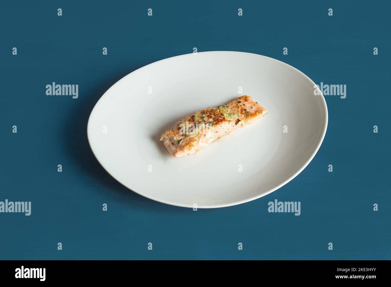 Servizio di un lombo di salmone al limone con prezzemolo e aglio su un piatto bianco e testurizzato sfondo blu studio. Pesce e cibo sano Foto Stock