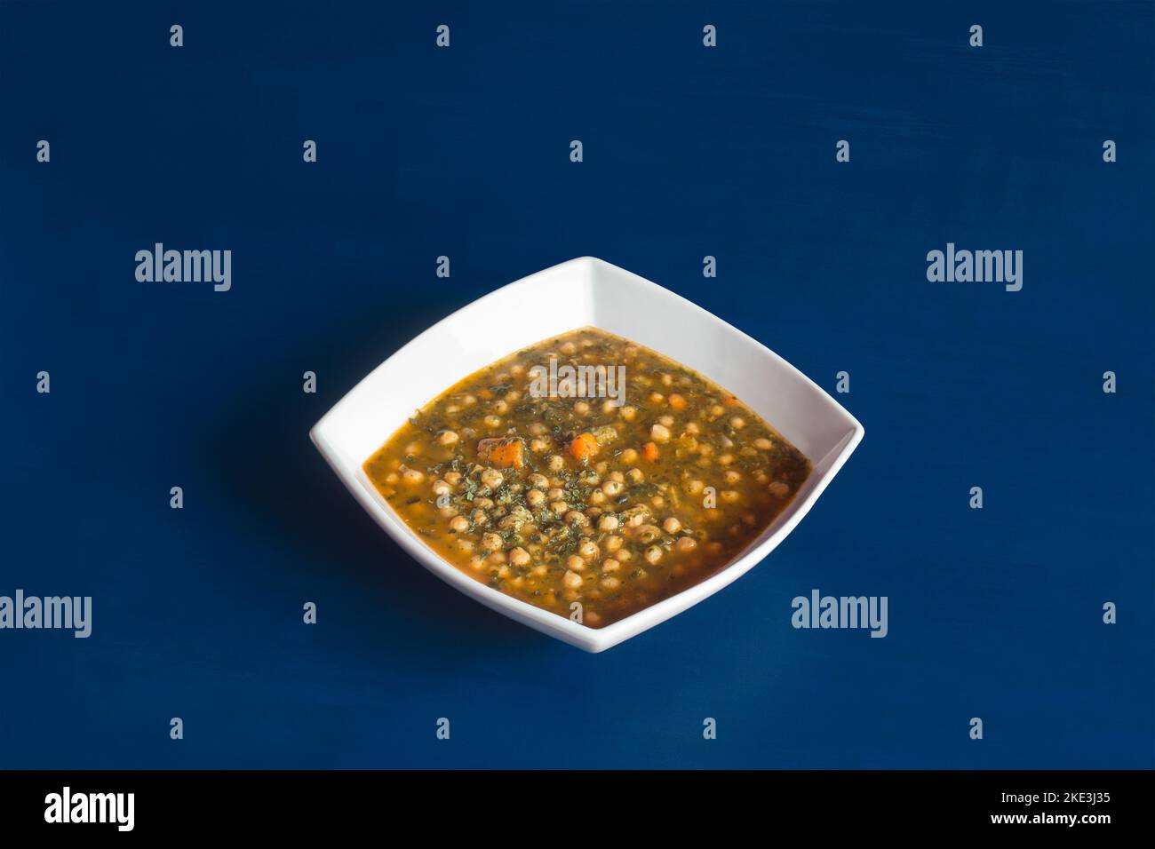 Un piatto vegetariano cotto di ceci con prezzemolo di spinaci e carote servito su un piatto di ceramica bianca a forma di diamante profondo su uno sfondo blu da studio Foto Stock