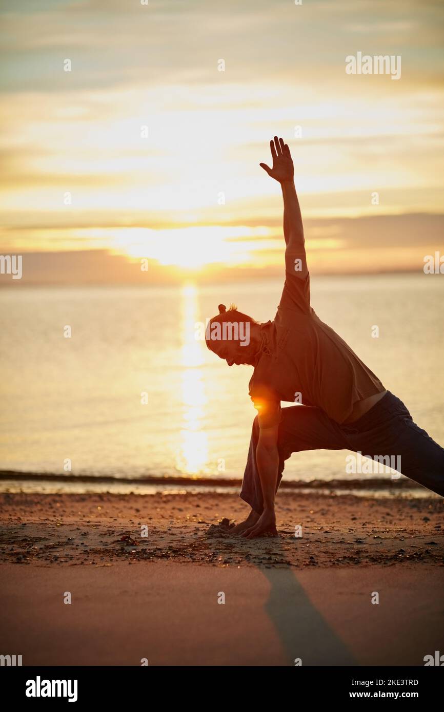 La natura è il partner ideale per lo yoga. Un uomo che pratica il triangolo posa durante la sua routine di yoga in spiaggia. Foto Stock
