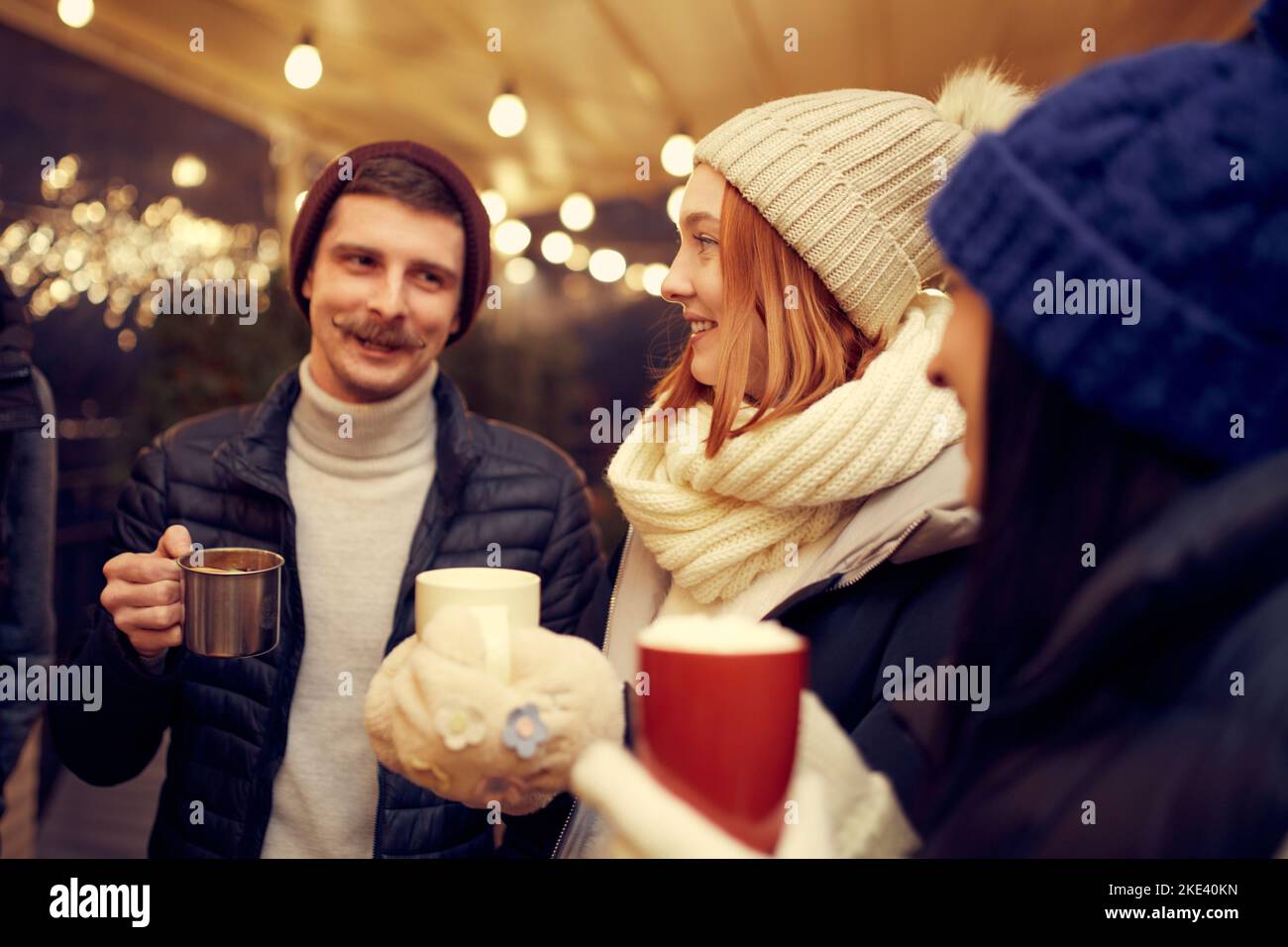 Gruppo di giovani che trascorrono del tempo insieme, bevendo VIN brulé, parlando, ridendo alla fiera invernale di sera. Vacanze invernali, vibrazioni natalizie Foto Stock