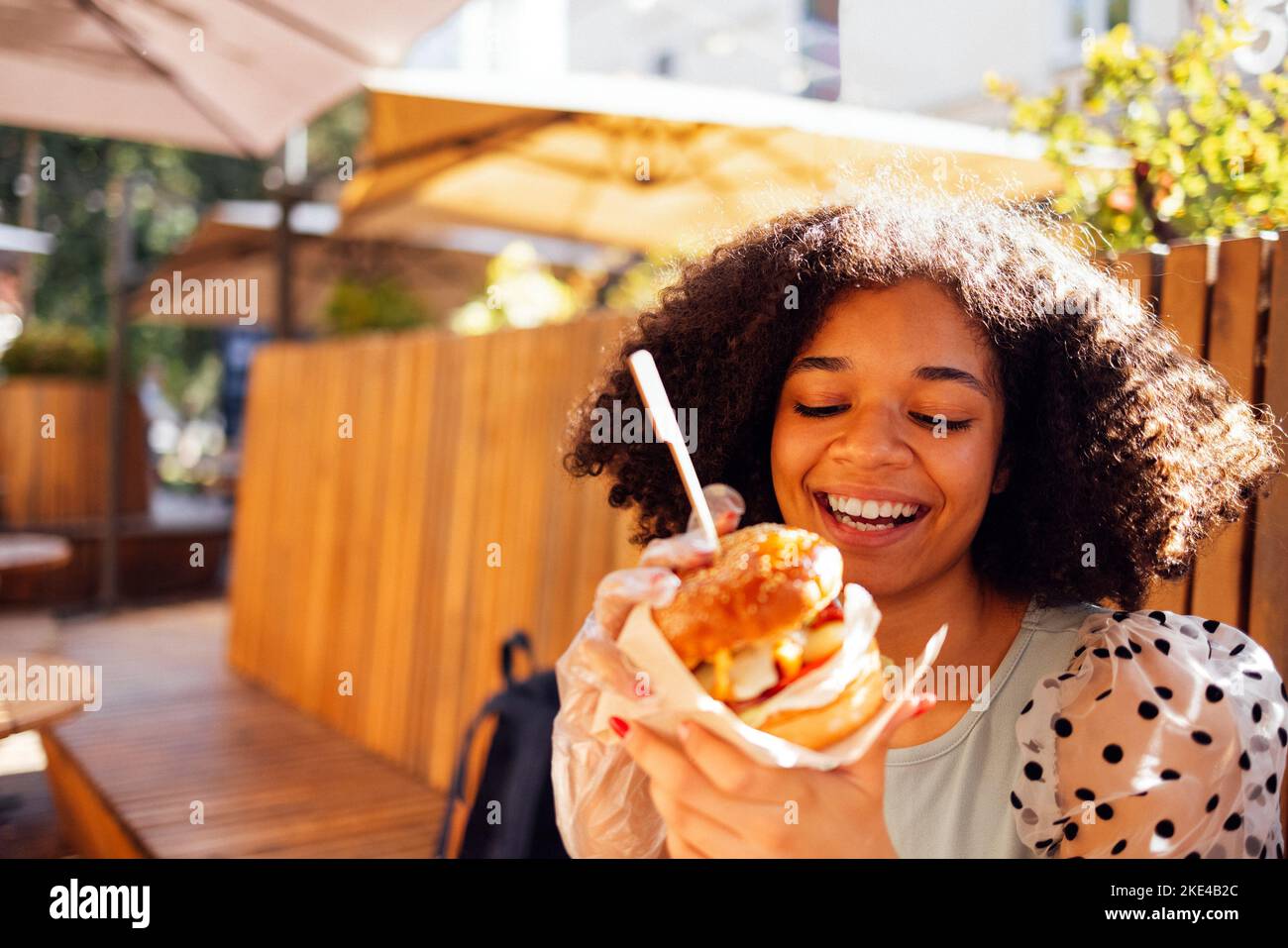 Giovane ragazza afroamericana che mangia hamburger classico e beve limonata in città. Donna outdoor indossa una blusa estiva con pois polka che mangia delic Foto Stock