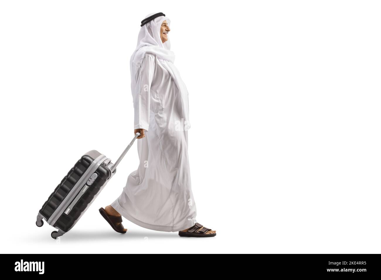 Profilo a tutta lunghezza girato di un uomo arabo in abiti etnici camminare e tirare valigia isolato su sfondo bianco Foto Stock