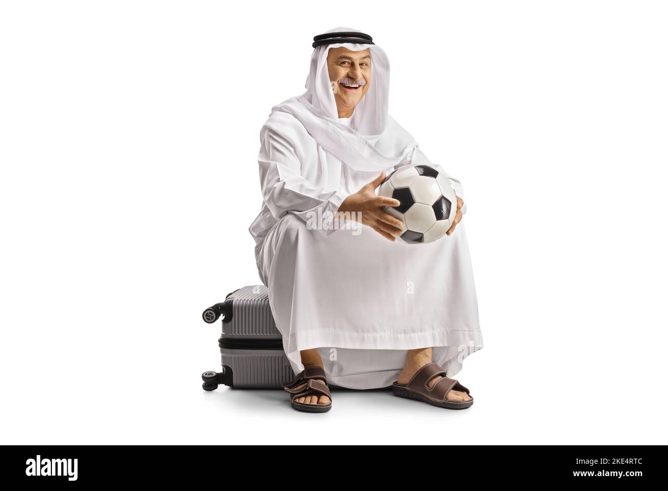 Uomo arabo maturo sorridente in abiti etnici seduto su una valigia e tenendo un calcio isolato su sfondo bianco Foto Stock