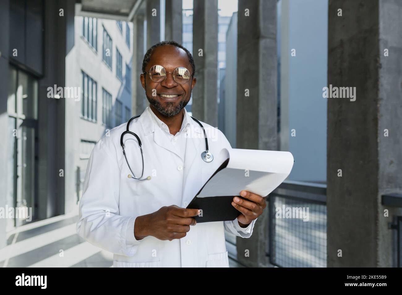 Ritratto di medico senior indossando occhiali afro-americani guardando la macchina fotografica e sorridendo al di fuori della clinica moderna, tenendo documenti del rapporto di trattamento del paziente, soddisfatto del risultato di lavoro. Foto Stock