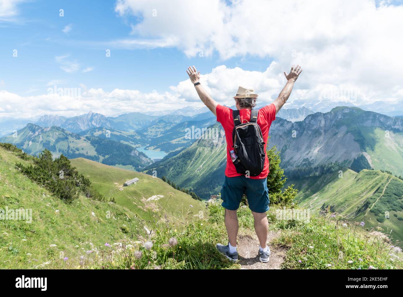 Uomo adulto anziano con cappello, zaino e camicia rossa con le braccia in alto guardando verso le Alpi svizzere. Foto Stock