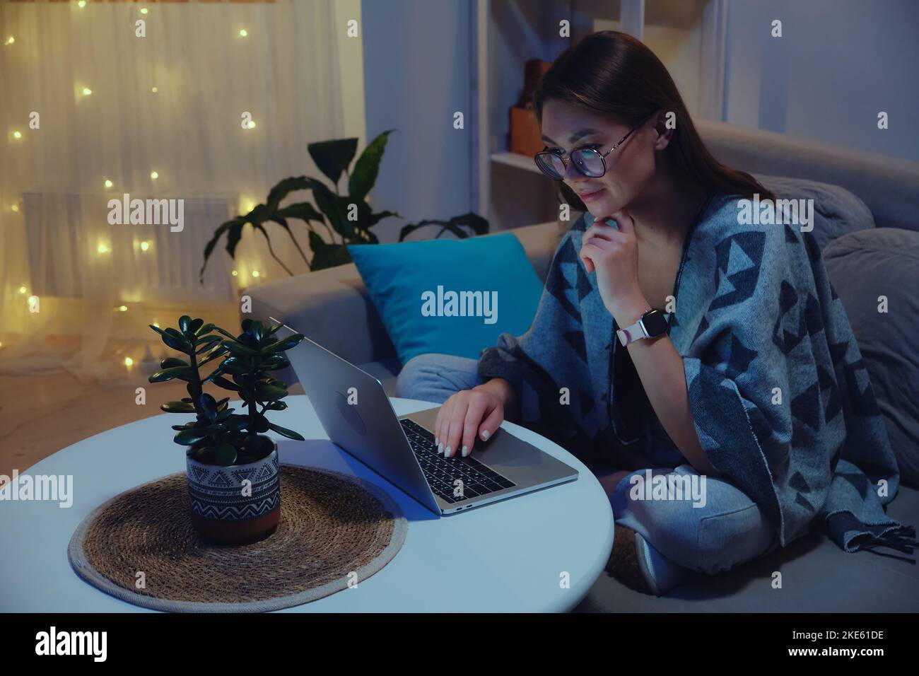 Giovane donna sorridente che usa il notebook di notte mentre lavora fuori orario con le luci di natale Foto Stock