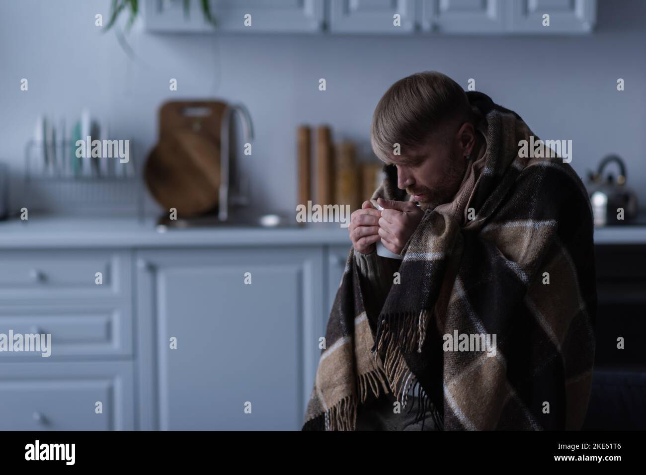 uomo congelato in coperta calda bere bevande calde durante il blackout di energia a casa Foto Stock