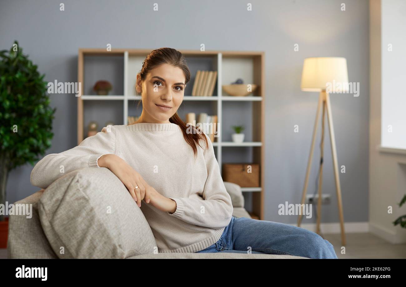 Ritratto di una giovane donna felice seduta sul divano a casa e guardando la macchina fotografica Foto Stock