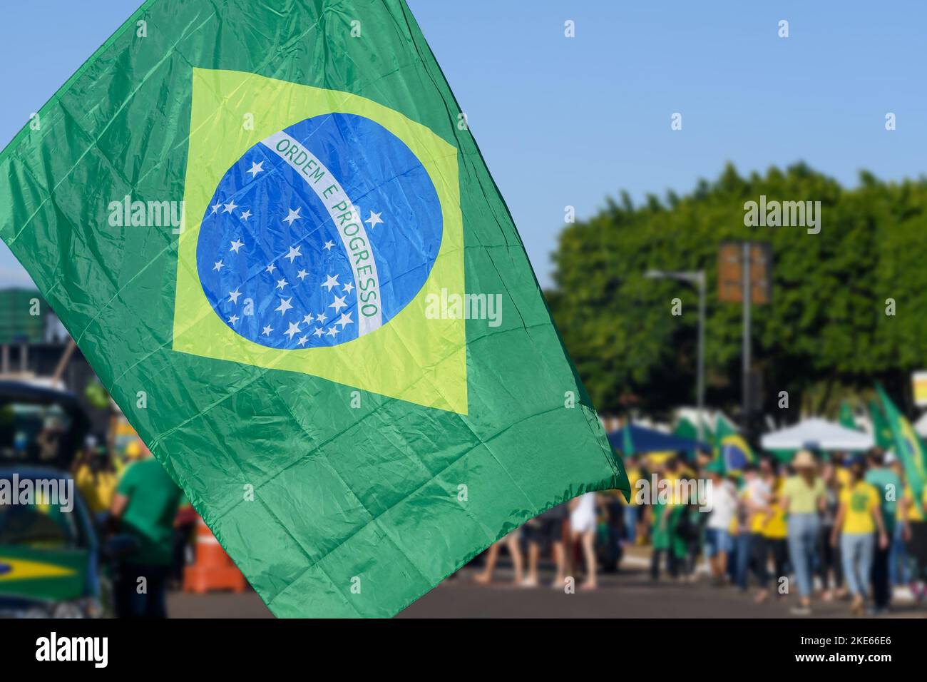 La bandiera brasiliana si è concentrata in primo piano e le persone brasiliane sulle strade sfocate sullo sfondo. Persone sfocate che indossano abiti verdi e gialli. C Foto Stock