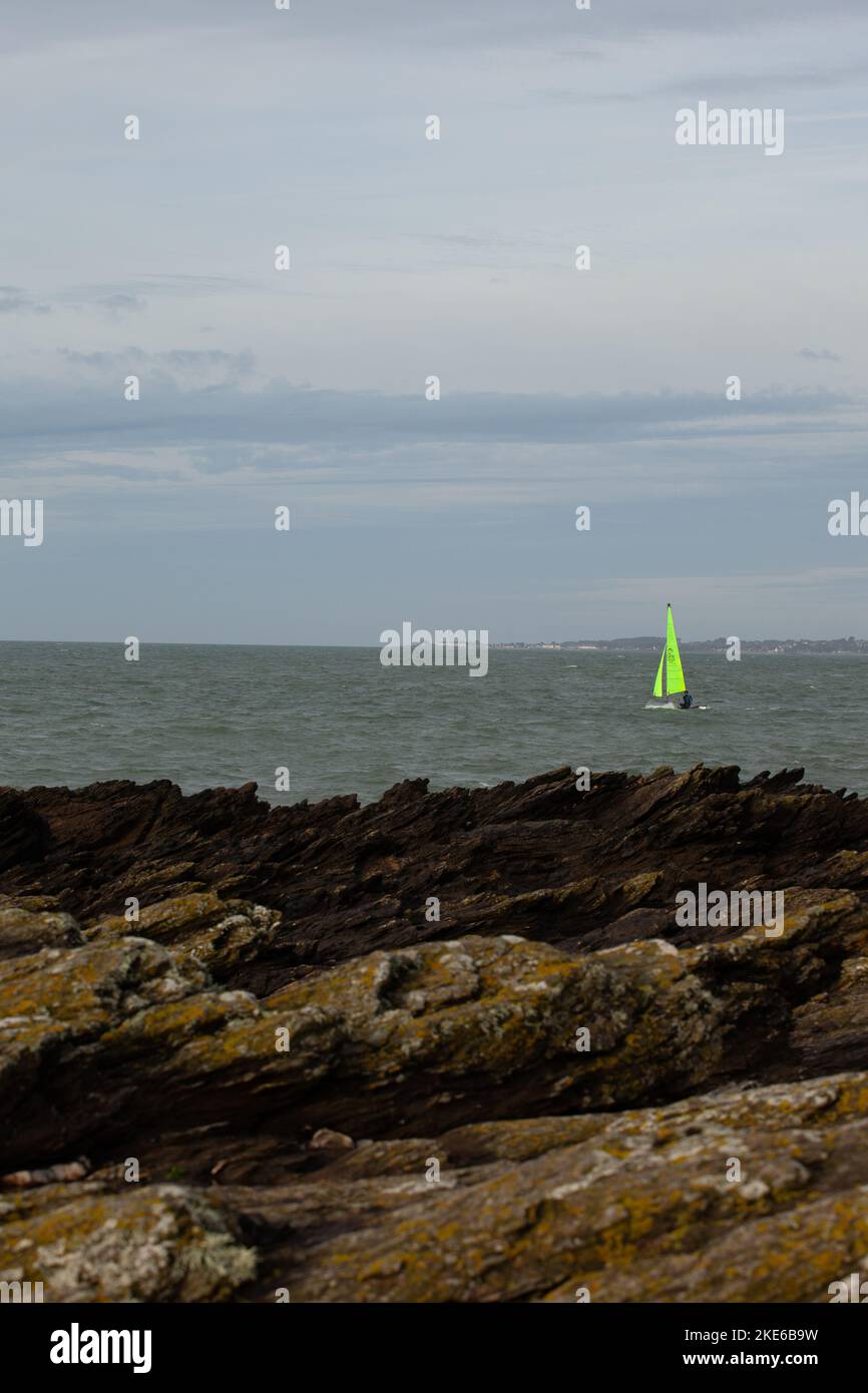Una barca a vela con una vela verde brillante naviga verso il mare oltre rocce taglienti. Foto Stock