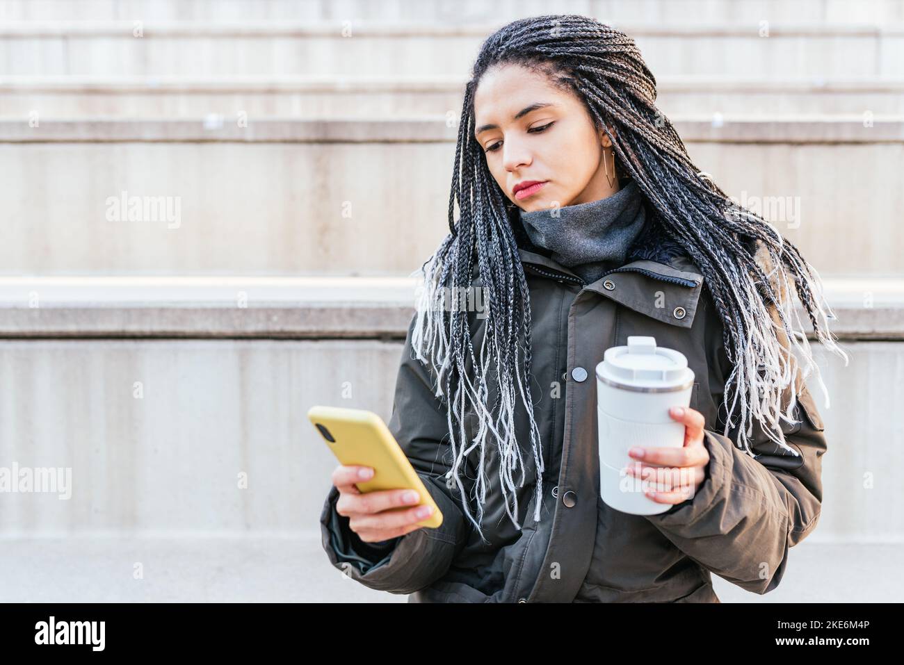 Donna ispanica focalizzata in outerwear con caffè caldo messaggi di testo sul cellulare mentre si siede sulla scala di strada della città Foto Stock