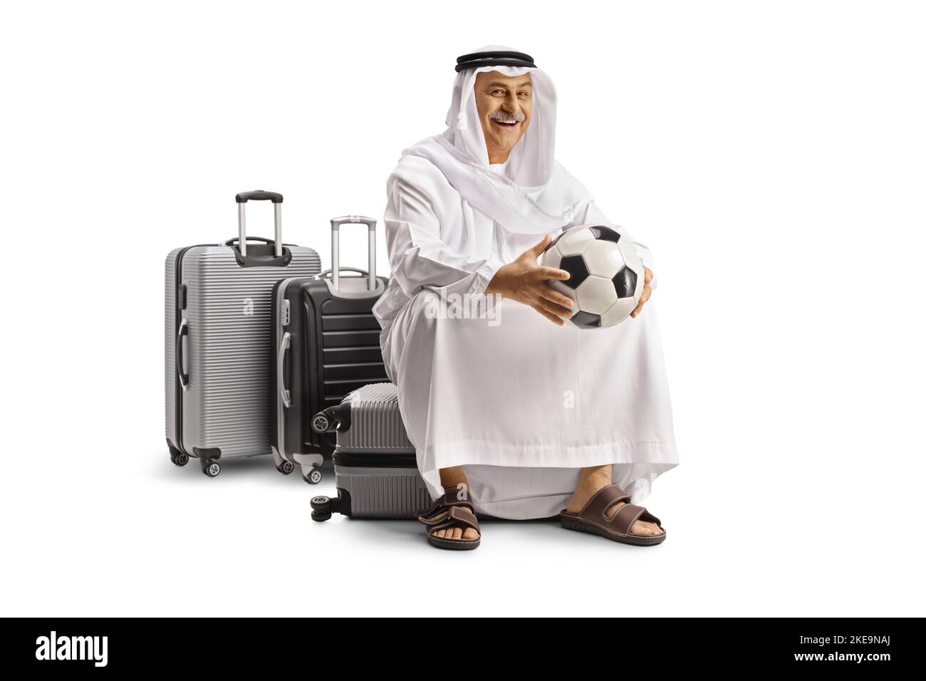 Sorridente passeggero arabo maturo in abiti etnici seduti su una valigia e tenendo un calcio isolato su sfondo bianco Foto Stock